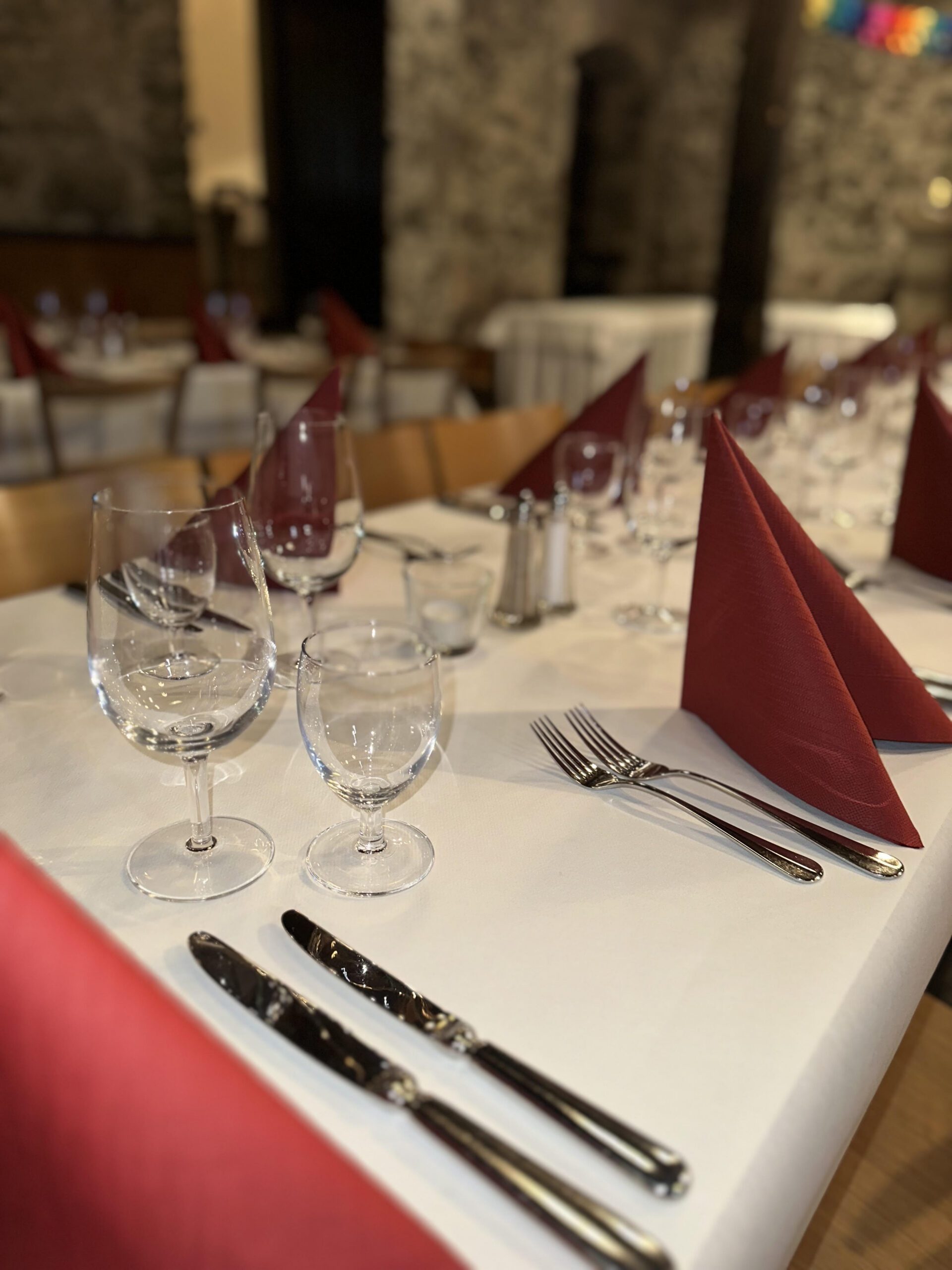 Eine Nahaufnahme eines ordentlich gedeckten Esstisches mit roten gefalteten Servietten, Weingläsern, Wassergläsern, Gabeln und Messern auf einer weißen Tischdecke in einem Restaurant mit Steinwänden.