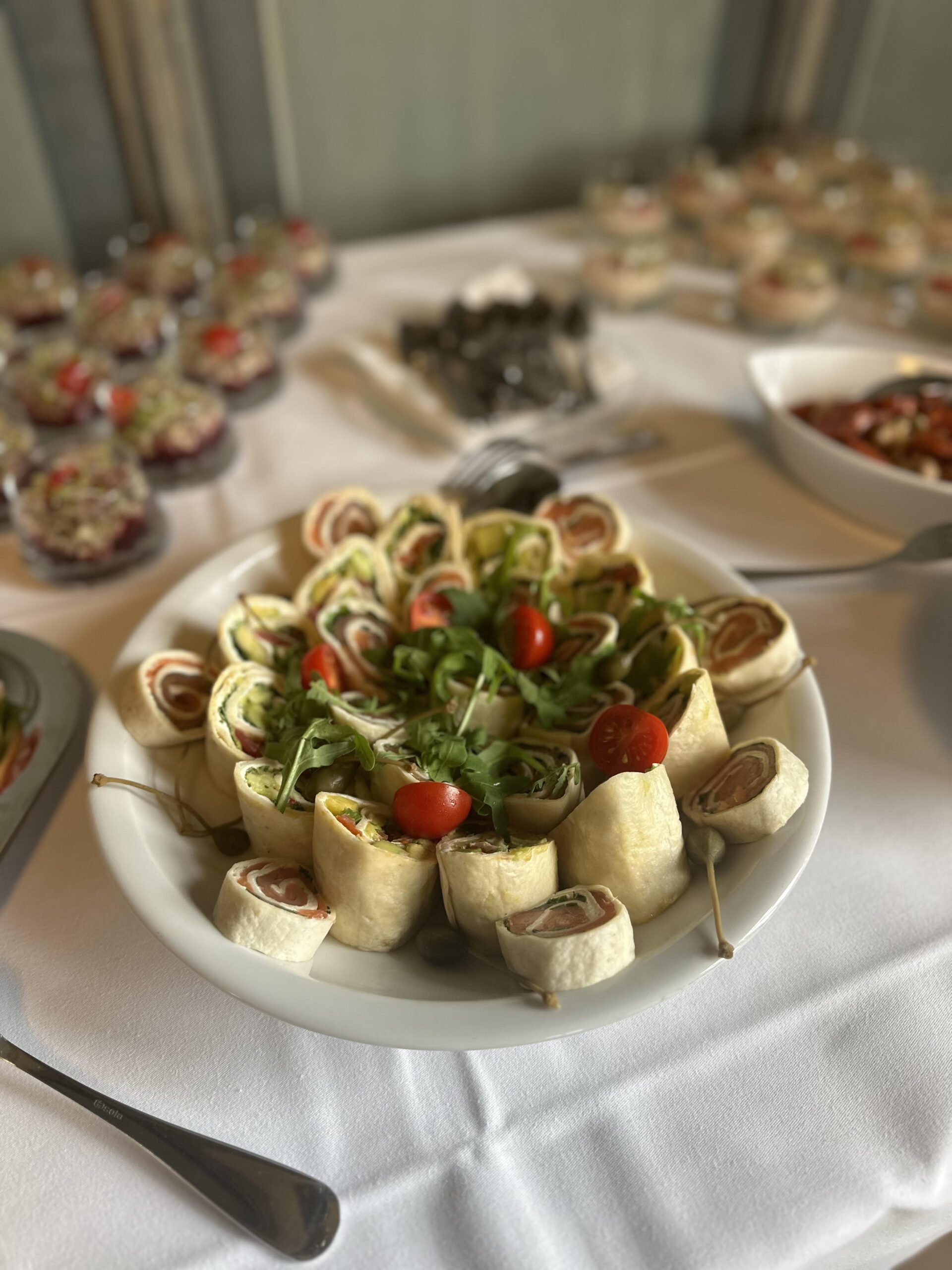 Ein Teller mit Tortilla-Wraps gefüllt mit Wurst, Gemüse und Käse, garniert mit Rucola, Kirschtomaten und Kapernbeeren, steht neben anderen verschiedenen Vorspeisen auf einer weißen Tischdecke.