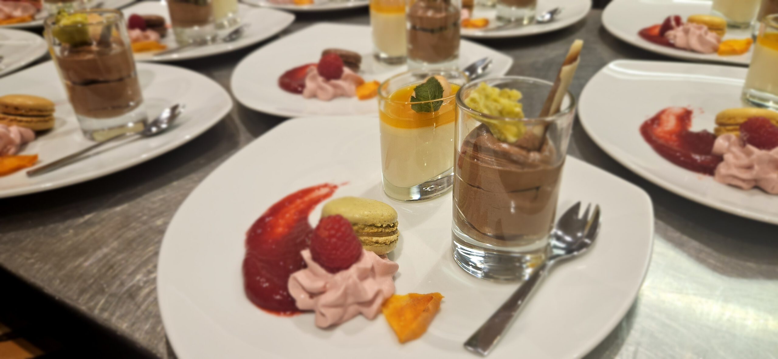 Eine Nahaufnahme eleganter Dessertteller mit Schokoladenmousse und Panna Cotta in Gläsern, Macarons, rosa Schlagsahne, Himbeeren, Fruchtsauce und Fruchtstücken, alles ordentlich auf weißen Tellern angerichtet.