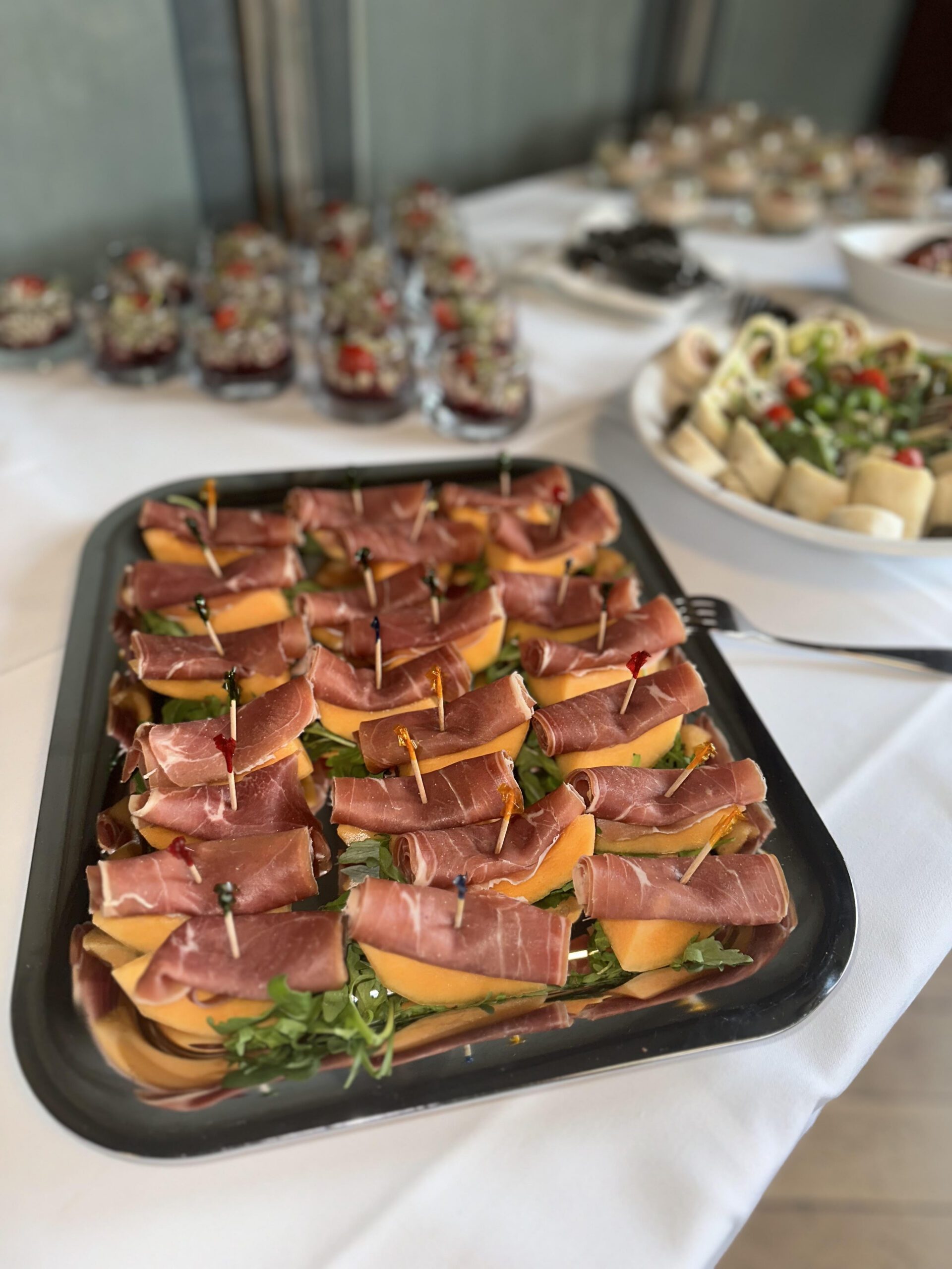 Auf einem Tisch steht ein Tablett mit in Prosciutto gewickelten Melonenscheiben und Zahnstochern, im Hintergrund sind weitere Vorspeisen und Salate zu sehen.