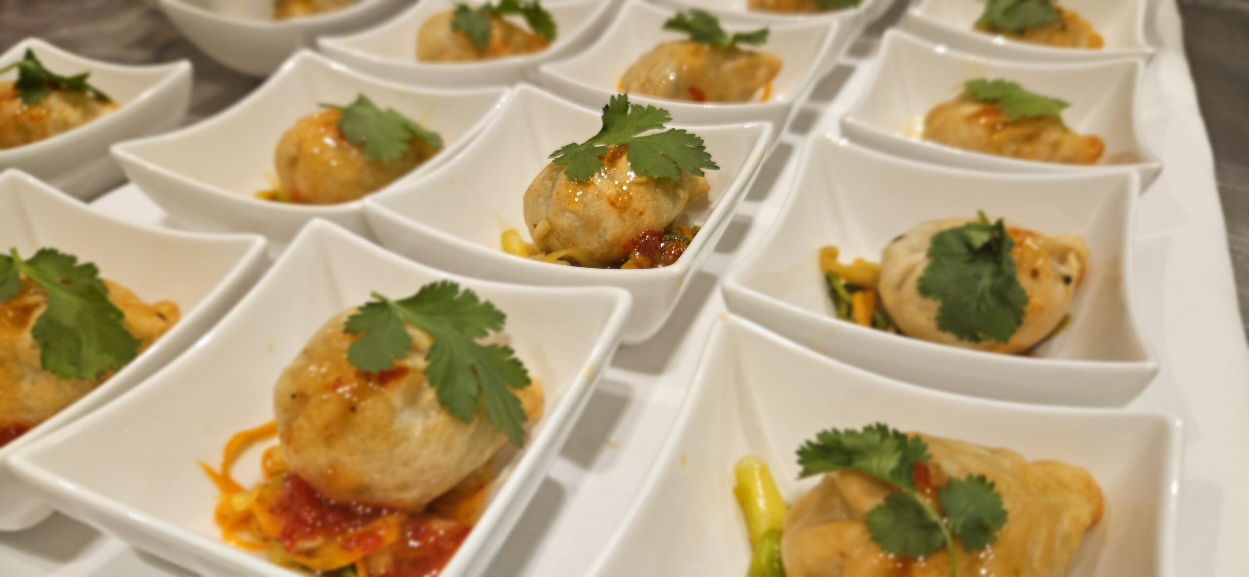 In Reihen weißer, quadratischer Schüsseln liegt jeweils ein mit Soße und einem frischen Korianderblatt garnierter Knödel, ordentlich zum Servieren auf einem Tablett angerichtet.