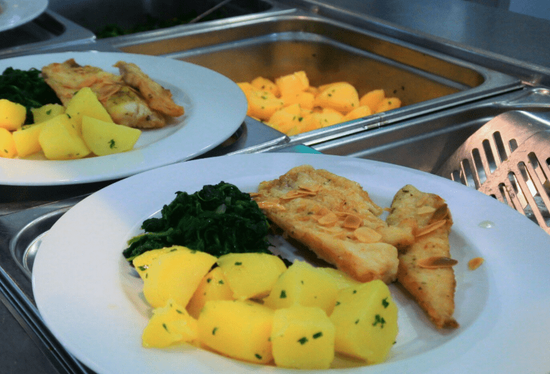 Zwei weiße Teller mit Portionen gekochter Fischfilets, garniert mit Mandeln, gedünstetem Spinat und gewürfelten Petersilienkartoffeln, auf einer Metalltheke neben einem Tablett mit weiteren Kartoffeln platziert.