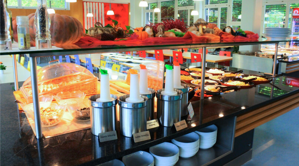 Eine hell erleuchtete Salatbar mit verschiedenen Toppings, Dressings in Spendern, Tellern, Schüsseln und Brotkörben. Im Hintergrund gibt es farbenfrohe Dekorationen und eine Auswahl an kalten Speisen.