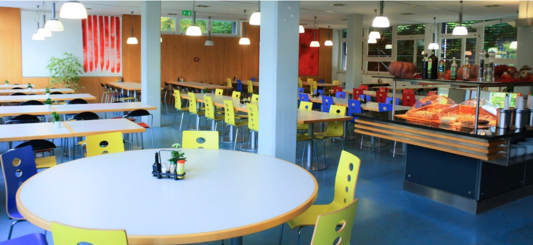 Eine hell erleuchtete Cafeteria mit gelben und blauen Stühlen an weißen Tischen, modernen Leuchten, einer Theke mit Speisenpräsentation und farbenfroher abstrakter Kunst an den Wänden. Große Fenster sorgen für Tageslicht.
